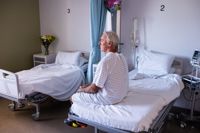 Elderly man sits on hospital bed looking out towards the ward.