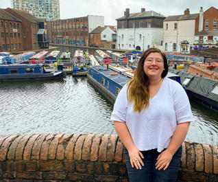 Lucy wears a white t-shirt and stands in front of canal boats.