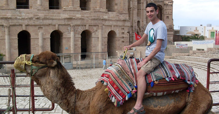Photo of CAHA alumnus Mark Desai (BA in Archaeology and Ancient History, 2014) astride a camel