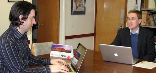 Photograph of two male academics in conversation at a table with laptops