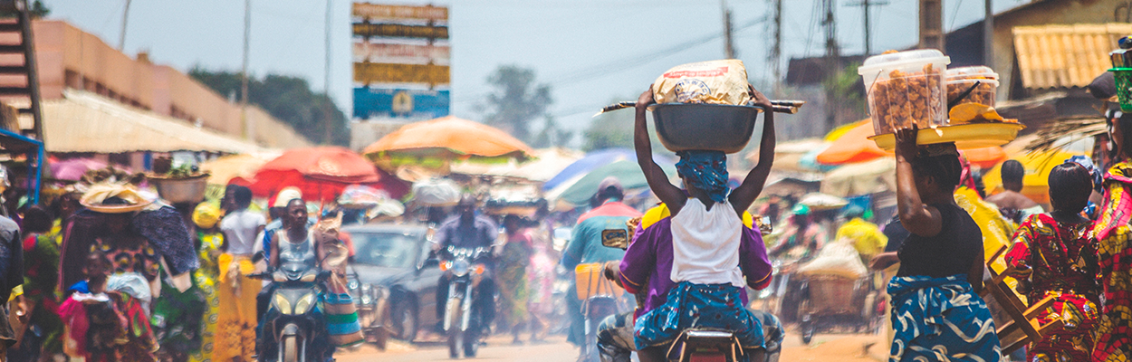 Image of a sub-Saharan African market as a backdrop to women carrying goods on their heads in a traditional way in the foreground