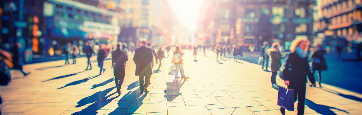 Blurred photograph of people walking on a large city center footpath, with the autumn sun shining through
