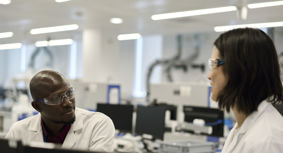 A male and female researcher wearing protective eyewear and a white lab coats, talking to each other