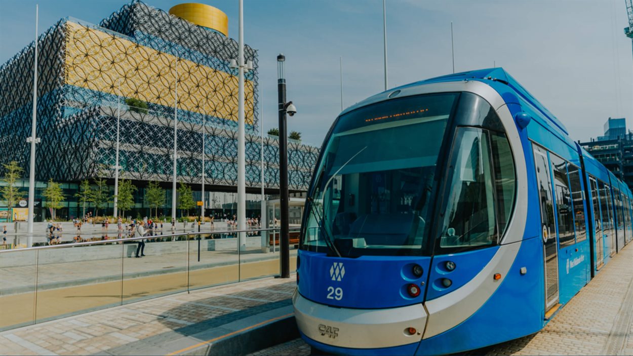 The front of a blue and grey tram is facing the camera. It is stopped outside a gold and blue building which is the Birmingham Central Library