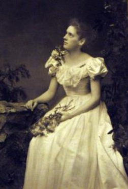 A sepia portrait of Ethel Chamberlain as a young woman, in an elegant white gown, holding flowers.
