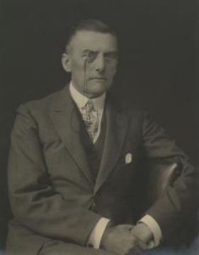 A portrait of Joseph Austen Chamberlain, leaning against a chair, wearing a suit and a monocle.