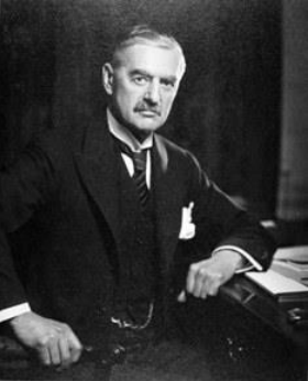 A portrait of a stern Neville Chamberlain leaning against a desk, wearing a dark suit, a wing collar, and a watch chain.