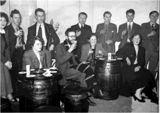 Black and white photo of happy male and female students sitting around barrels drinking.