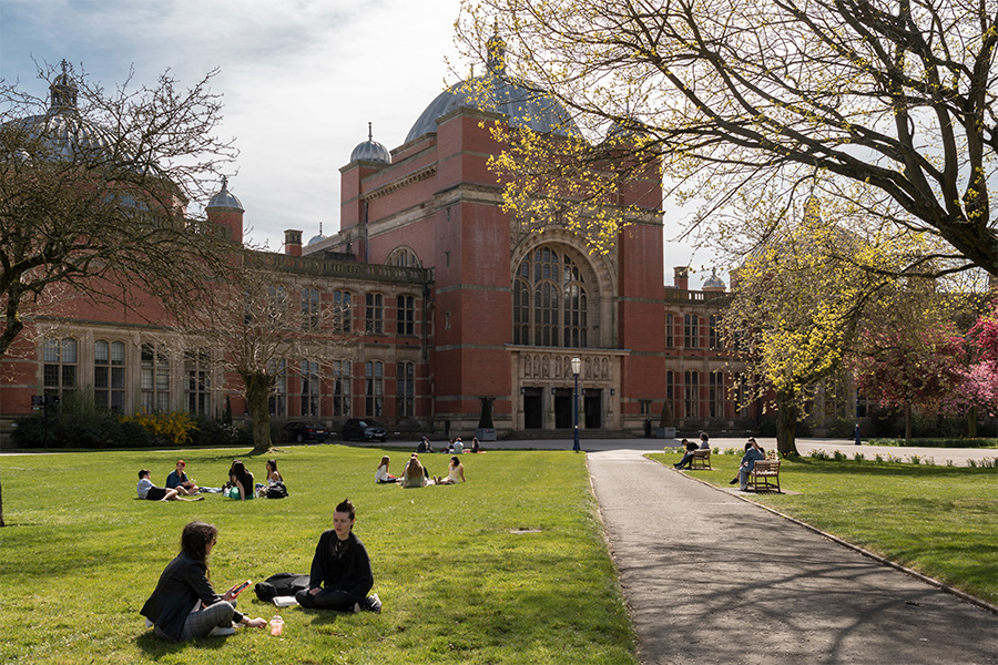 Students sitting on the grass in front of the redbrick buildings of University of Birmingham's Aston Webb building
