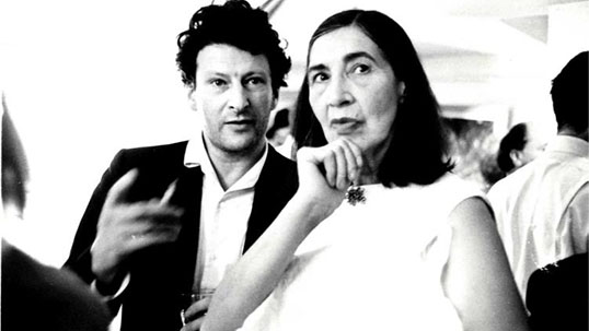 Black and white photograph of Kathleen Garman and Lucian Freud at an event – Freud is wearing a suit jacket and with white open-necked shirt and holds a full glass and a cigarette. He appears to lean in towards Garman. Both look towards the photographer