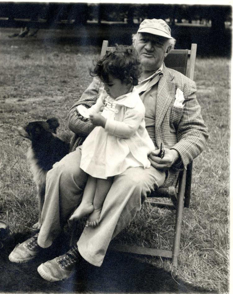 Black and white photograph of Epstein dressed casually in a baseball cap, sitting on a deckchair outside on the grass. A small child of about two, sits on his lap, seemingly captivated by an object in her hands. A dogs sits by Epstein’s chair and looks aw