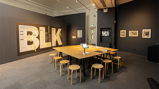 A photograph of a gallery space. The walls are painted dark grey and the floor is carpeted. On one wall there are three framed posters and a screen. The other wall has a timeline placed in the letters BLK in white font with yellow sticky notes. In the centre of the room is a large square wooden table surrounded by wooden stools. The table has a screen, a folder, leaflets, and other pieces of paper on it.