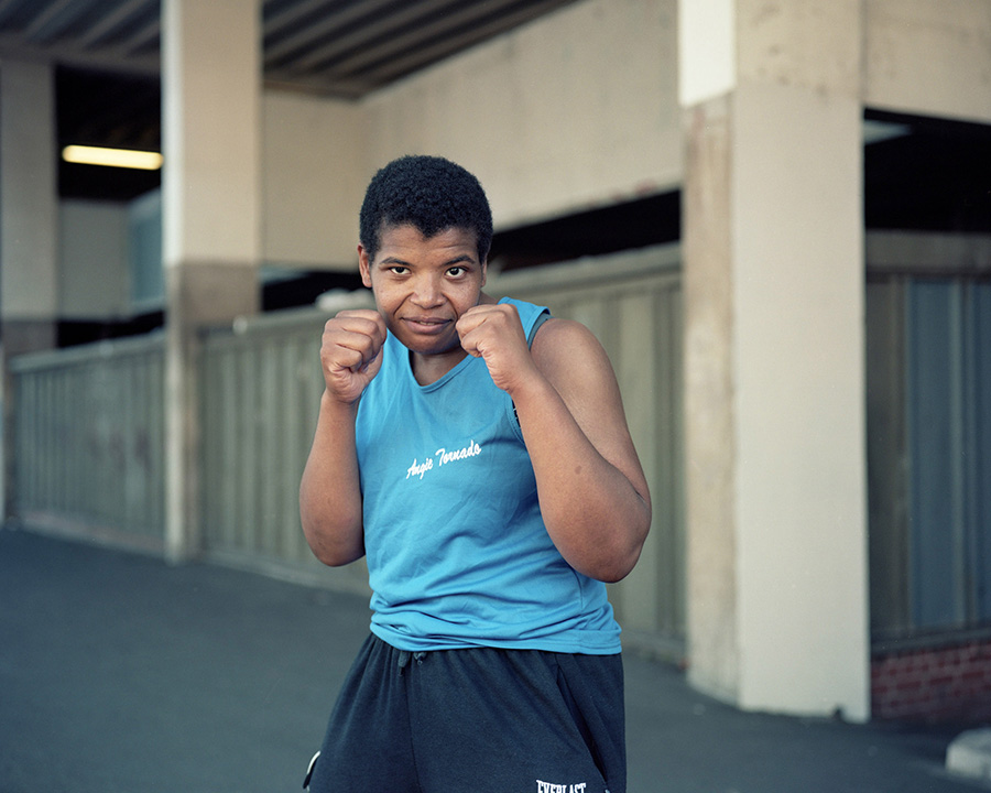 A woman of colour posing with her hands held in front of her in an agile boxing position. She is not wearing boxing gloves. She stands in front of a building.