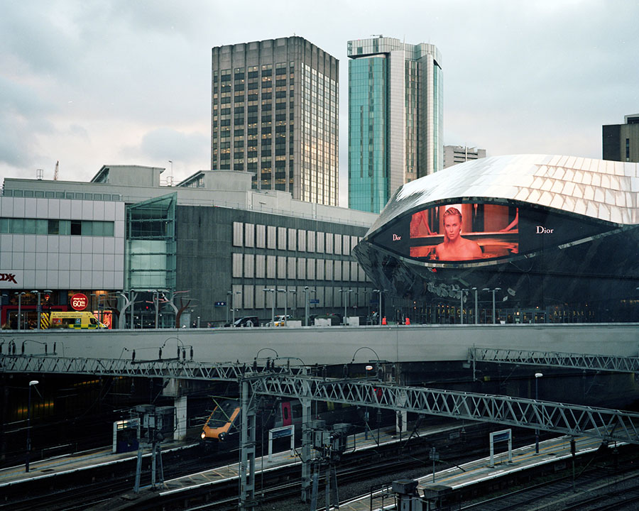 New Street Station tracks framed by various high-rise buildings. The front of New Street with the large screen is also visible.