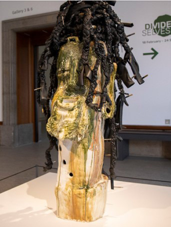A large sculpture of a woman’s head and neck. The face is contorted, with closed eyes and a gaping mouth, and there are ivy-green drips down the face and neck. The hair is made from braided tyres and is held up with metal pins and tubes.