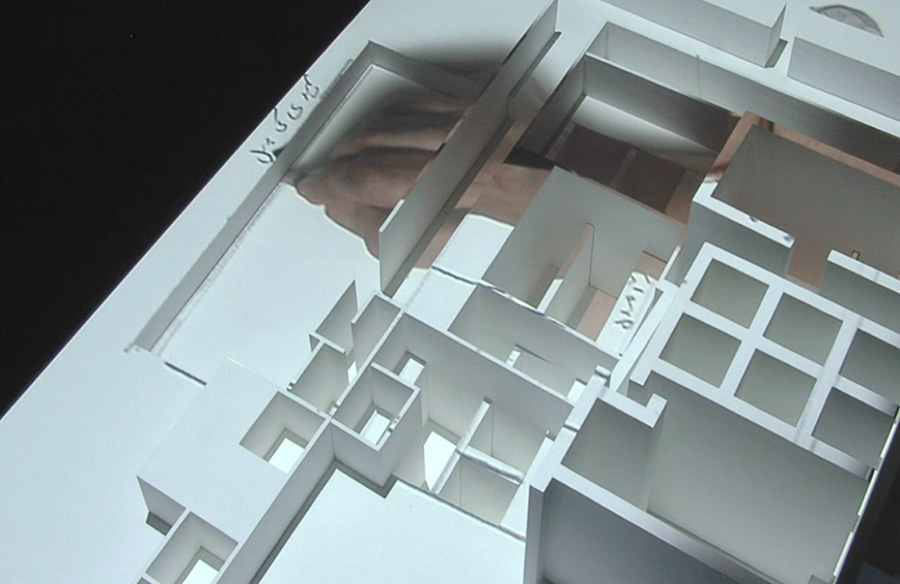 A small white paper model of a building. Onto it, an image of a hand writing in a foreign language is projected.
