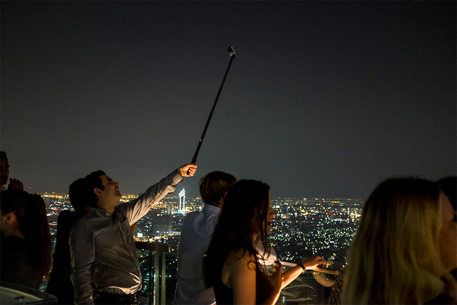 A night time scene at the Sky Bar in Belua State - a man is holding a selfie stick to take a picture of himself while he looks across the illuminated city