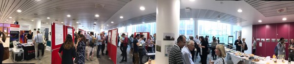 Panoramic view of a group of people in a wide open space room where some people are talking to each other and others are reading posters on red poster boards.