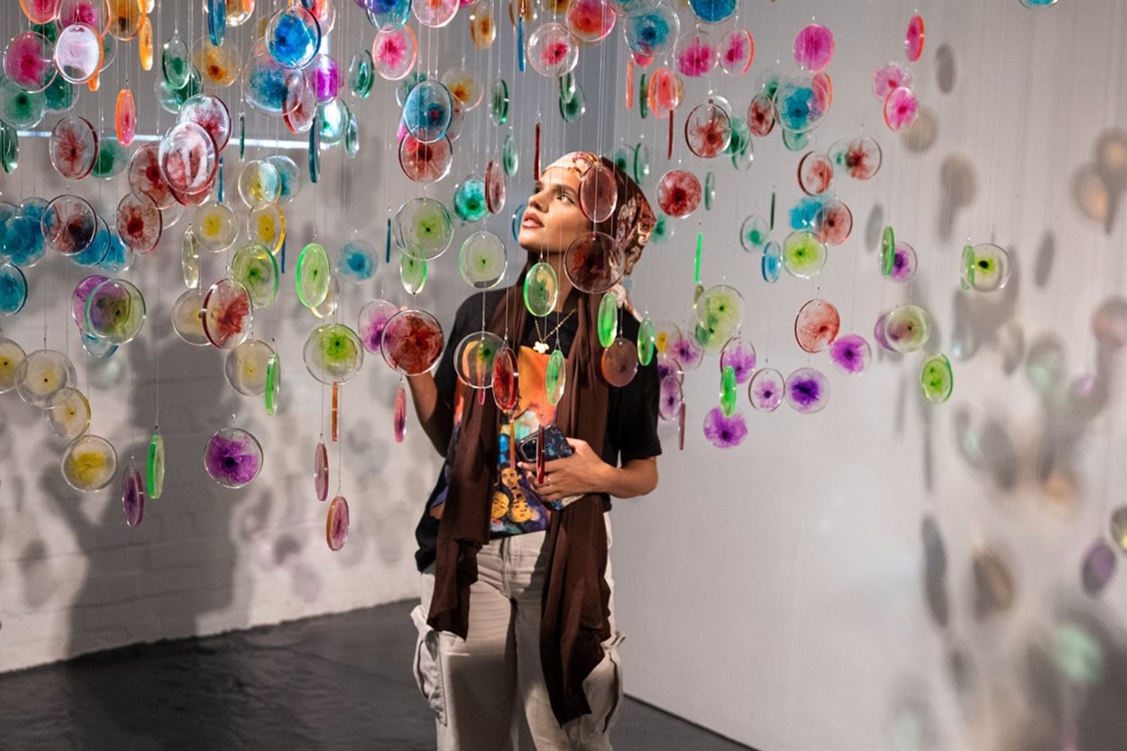 The photograph shows a young South-Asian lady in her twenties wearing a headscarf and admiring a hanging artwork which she is reaching out to touch in an art gallery space. The artwork consists of hundreds of bright and multicoloured discs. These discs artistically represent cells and the flow cytometry process within science which involves dyeing different cells and cell components into different colours to study different features. In this case the artwork references a project looking at residual leukaemia cells in disease monitoring after treatment.
