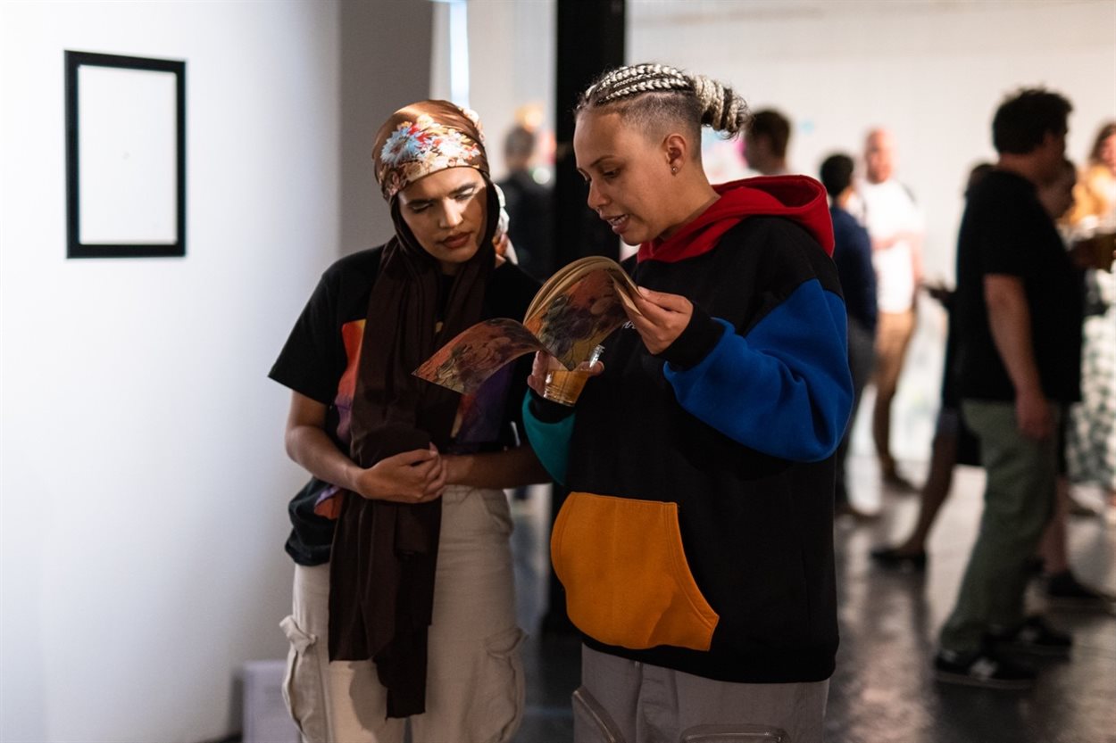 The photograph shows two young women in their twenties, one of South-Asian descent and another or Black or Afro-Carribean descent. The two are pictured within an art gallery space reading over a booklet together which features the interpretation text relating to the different artworks on display. In the background are several other men and women looking at the artworks featured throughout the room.