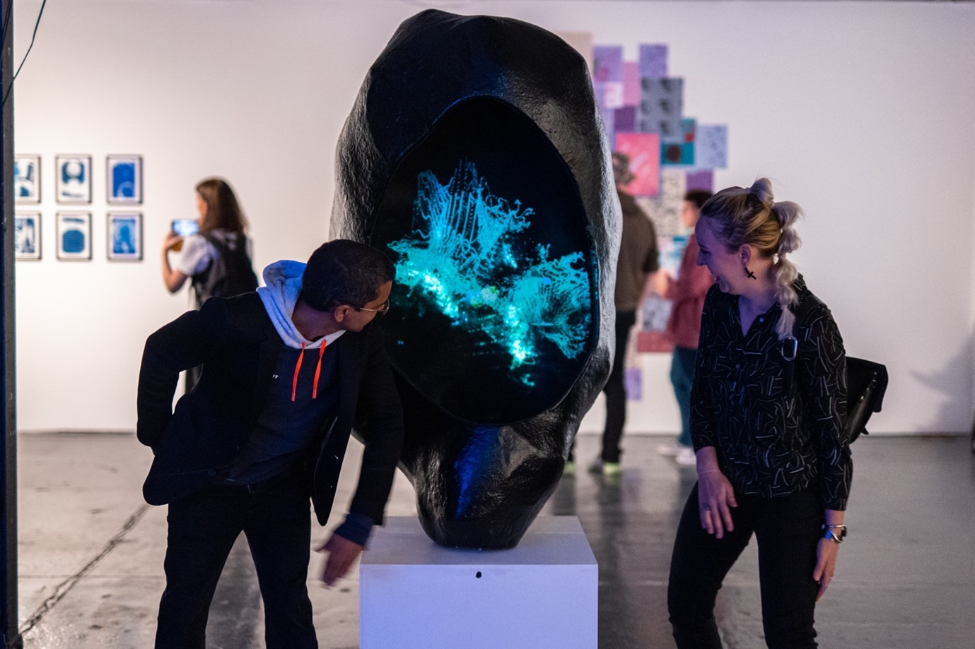The photograph shows a blond, white female and a south-asian male interacting with a motion sensitive artwork within an art gallery space. The artwork consists of a large black shell-like structure into which is projected a colourful turquoise audio-visual display of an abstract matrix which shifts in shape and which artistically represents the process of DNA methylation.