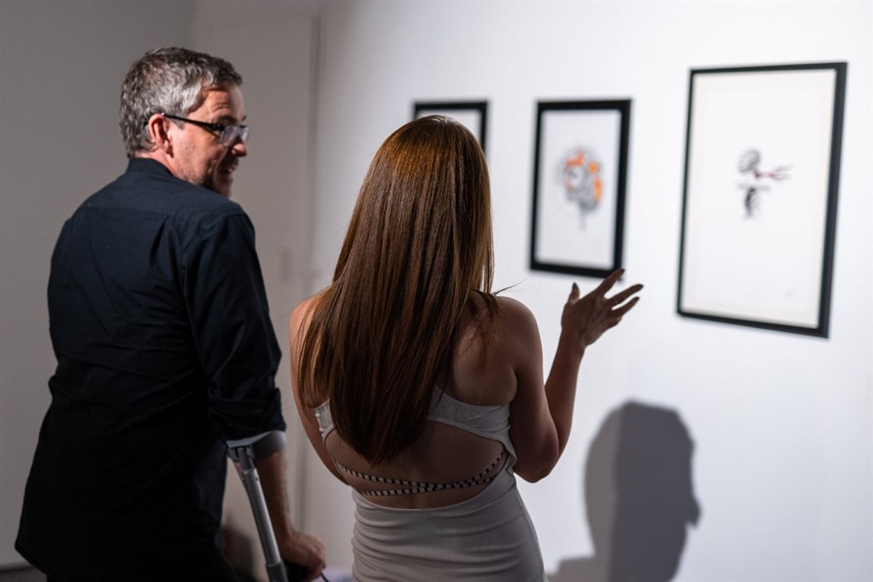 The photograph shows a white man in his forties using crutches and a brunette woman in her thirties discussing a series of drawings on display on the art gallery wall. The drawings which feature different depictions of the brain tell an abstract visual story about coming to terms with an epilepsy diagnosis and the emotions felt by the artist.