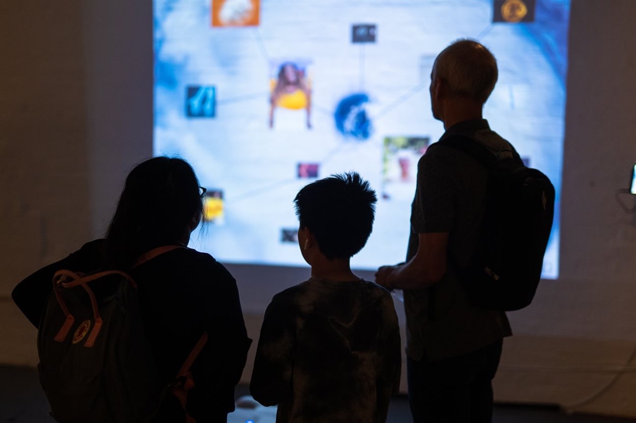 The photograph shows the outline of a family of three consisting of a mum, dad and young son around 10 years of age in an art gallery setting. The family are looking away from the camera and viewing a digital artwork projected on to the wall in front of them. This colourful projection which appears blurry in the photo includes a network of different pictures including photos of two women and natural events such as storms and lightning. The artwork using metaphor to depict and connect the emotions of people with acromegaly and visitors can listen to audio recordings that explain the meaning behind each image featured in the artwork.