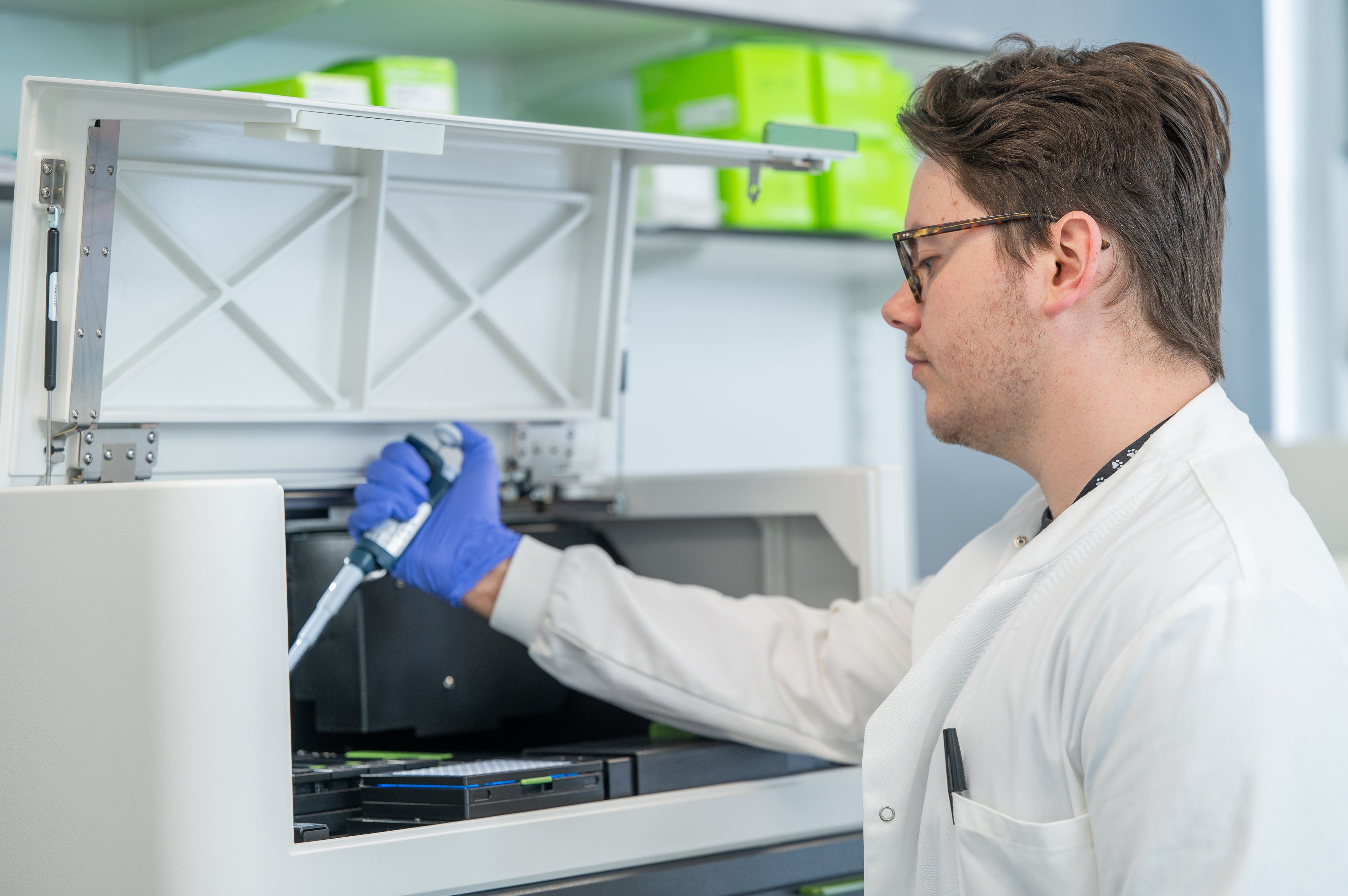 Male scientist wearing a white lab coat and blue rubber gloves holding a pipette in a lab setting.