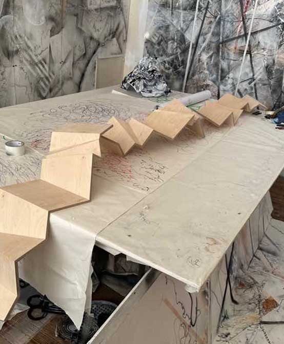 Art studio with sketches covering the walls, and a long wooden structure (representing a zig-zag shape) on a table