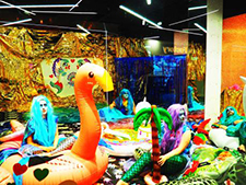 An image of Alex’s exhibition with inflatable palm trees and animals and people dressed as mermaids.