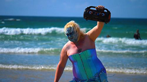 An image of Alex dressed as a mermaid on the beach holding a stereo.