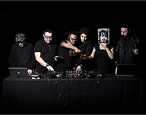Six men wearing black standing behind a DJ deck.