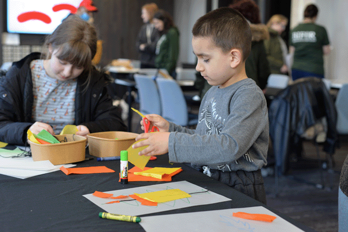 Two young children having fun doing arts and crafts at a table covered in coloured paper, glue, scissors and other crafting items, with more children and adults doing the same in the background.