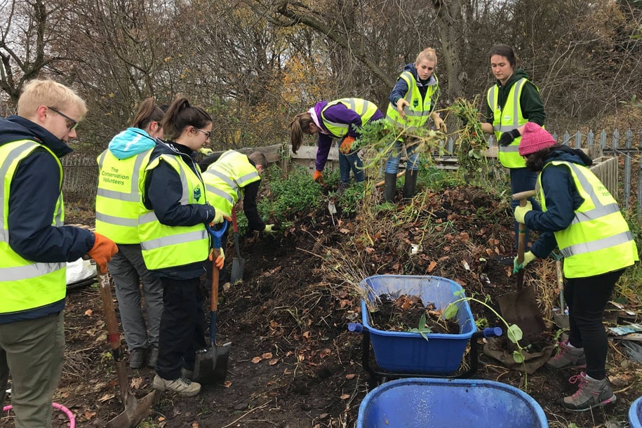 A group of student volunteers wearing high-visibility vests clearing foliage from a canal towpath.
