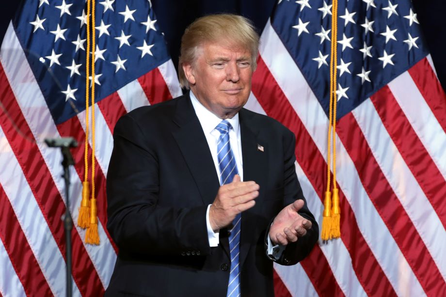President Donald Trump applauding, standing in front of two American flags.