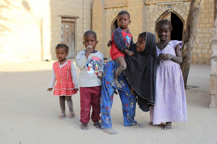 Five Malian children standing together on the dusty street of a town.