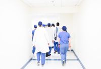 A group of doctors in white coats and medical scrubs walking down a bright, white corridor.