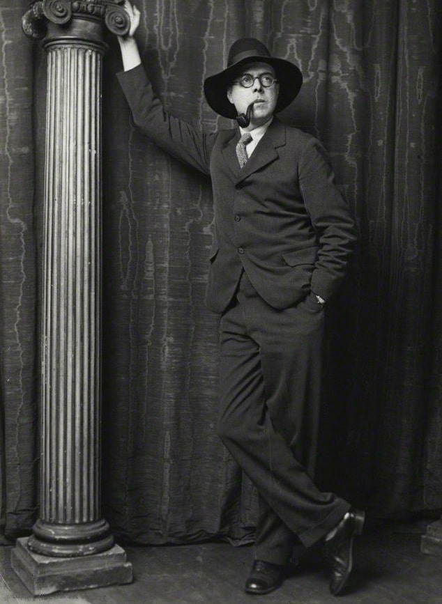 Wyndham Lewis wearing a suit and smoking a pipe, leaning against studio prop pillar