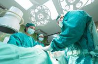Three surgeons in surgical gowns and face masks work on a patient under the lights of an operating theatre.
