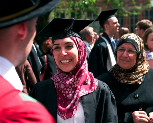 A young woman in her graduation gown smiling chatting to her lecturer, with a proud mother looking on in the background.