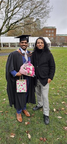 Naren and Jeevan standing together on campus on Naren's graduation day. Naren is wearing cap and gown.Both are smiling and Naren is holding a bunch of pink and white flowers, and a gift bag.