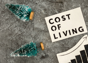 Miniature Christmas Trees on a grey background next to a label reading "cost of living" and a cut out graph with an upwards trajectory.