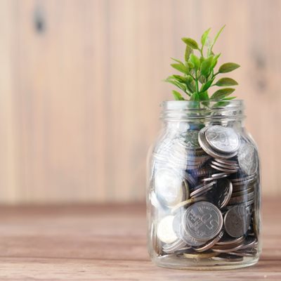 A jar of coins modelled to look like a plant pot with a green shoot growing from the top sits in front of a wooden background