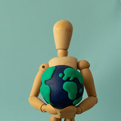 A wooden artist's figure holds a plasticine world in its hands, set against a blue-green background.