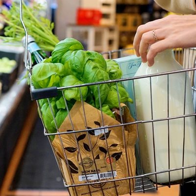 A metal shopping basket being carried by an eco-conscious shopper. In the basket are loose vegetables, a paper packet and a glass bottle of milk, no plastic to be seen.