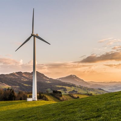 A wind turbine sits in a green field at either sunrise or sunset. There are mountains in the background. It's a serene image.