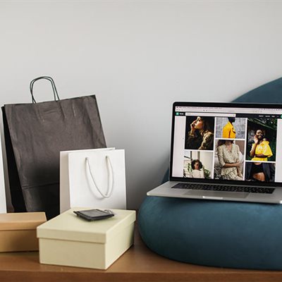 A group of multi-coloured paper shopping bags sit next to a laptop on a blue sofa cushion. The laptop displays images of fashion pieces.