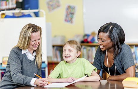 two female teachers in a classroom assisting a young boy with SEN with his reading