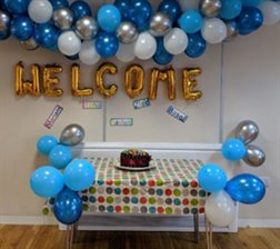 a table with a cake and balloons on it with a welcome sign above it