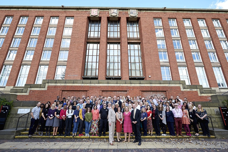 A large group of librarians stand on the steps outside the old Main Library: a tall red-brick 1950s building with the university's crests high above the doors.
